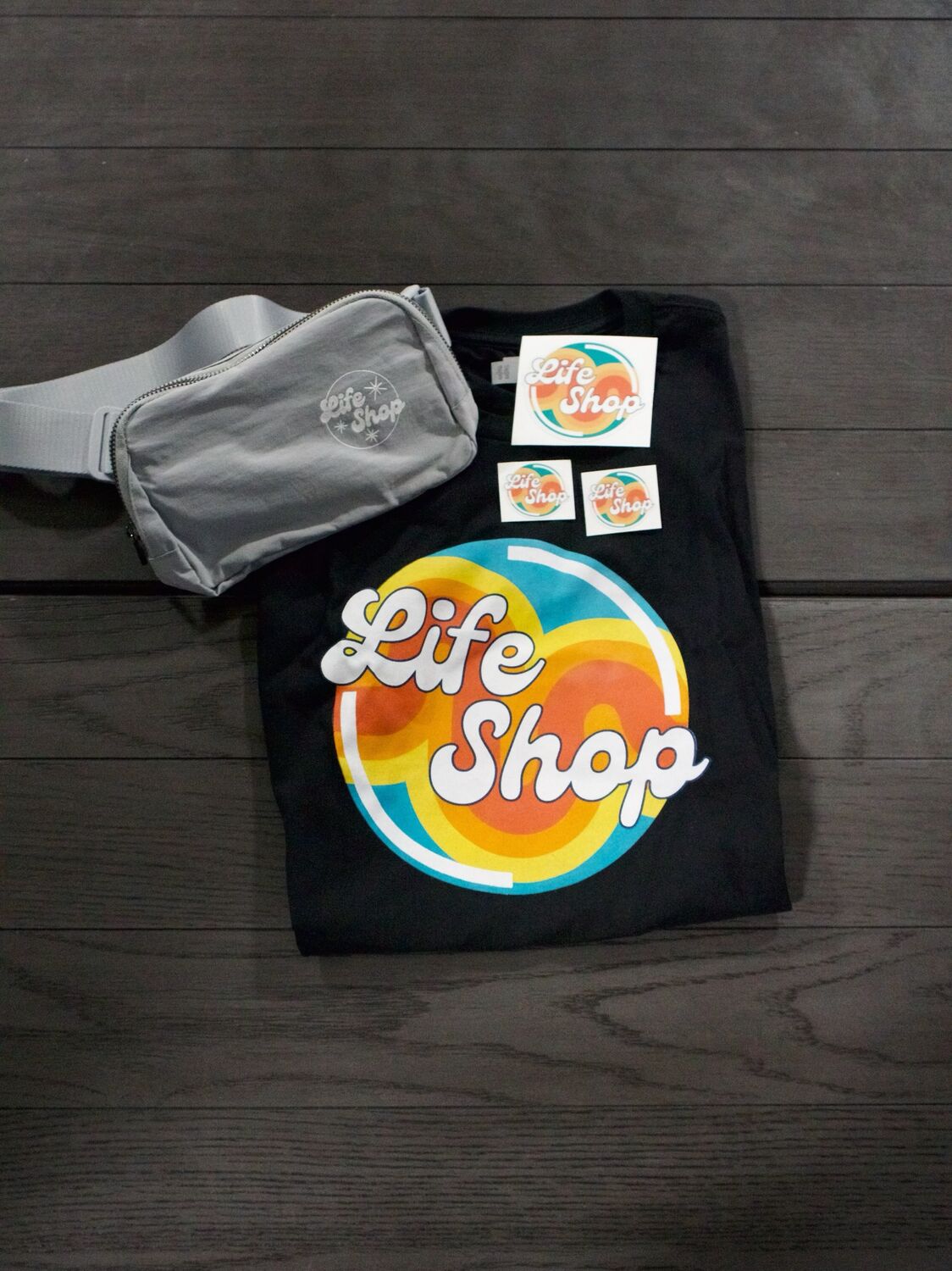 A folded black Life Shop tee, a grey belt bag and Life Shop stickers laid out together