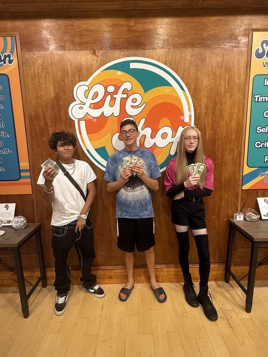 Three teens holding their winnings in front of the Life Shop logo wall